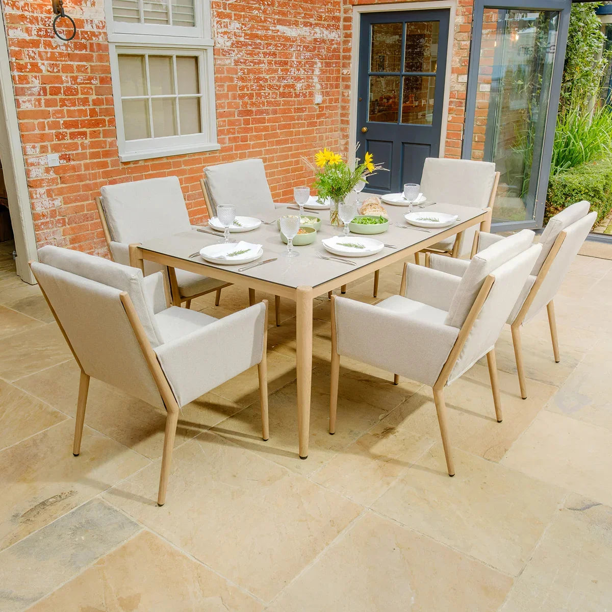 Beige outdoor dining set with eight cushioned chairs on stone patio, table set for a meal