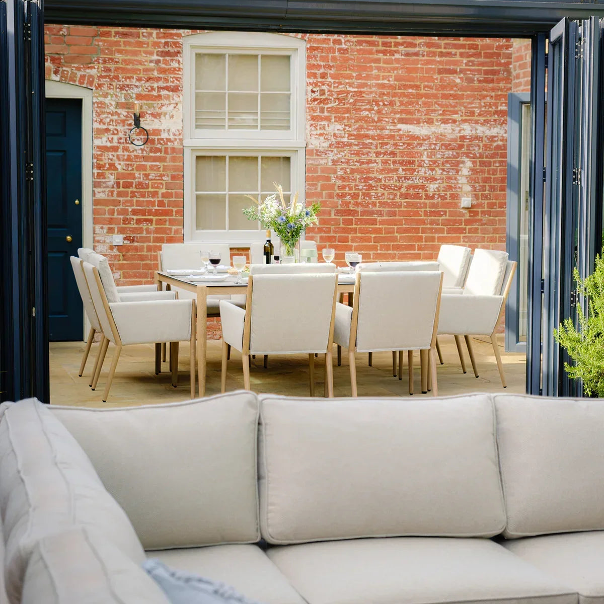 Outdoor dining table with eight cream cushioned chairs on patio against brick wall