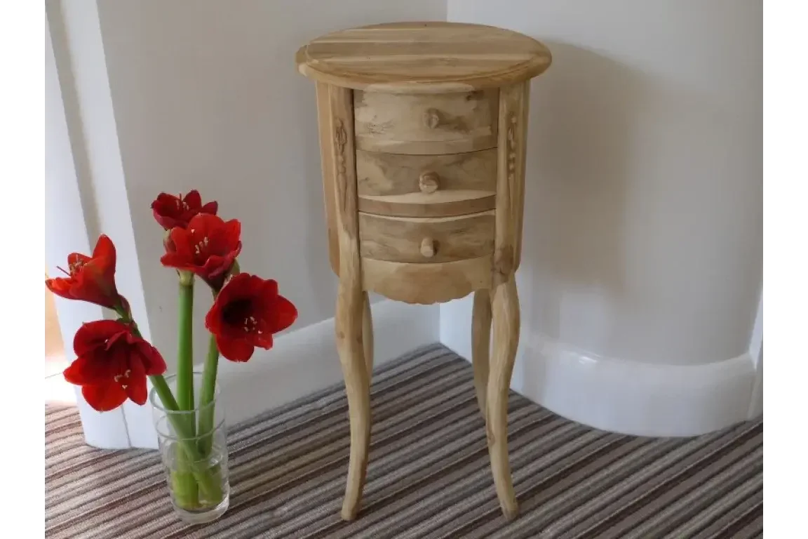 Round unfinished wooden bedside table with three drawers beside red flowers in glass vase
