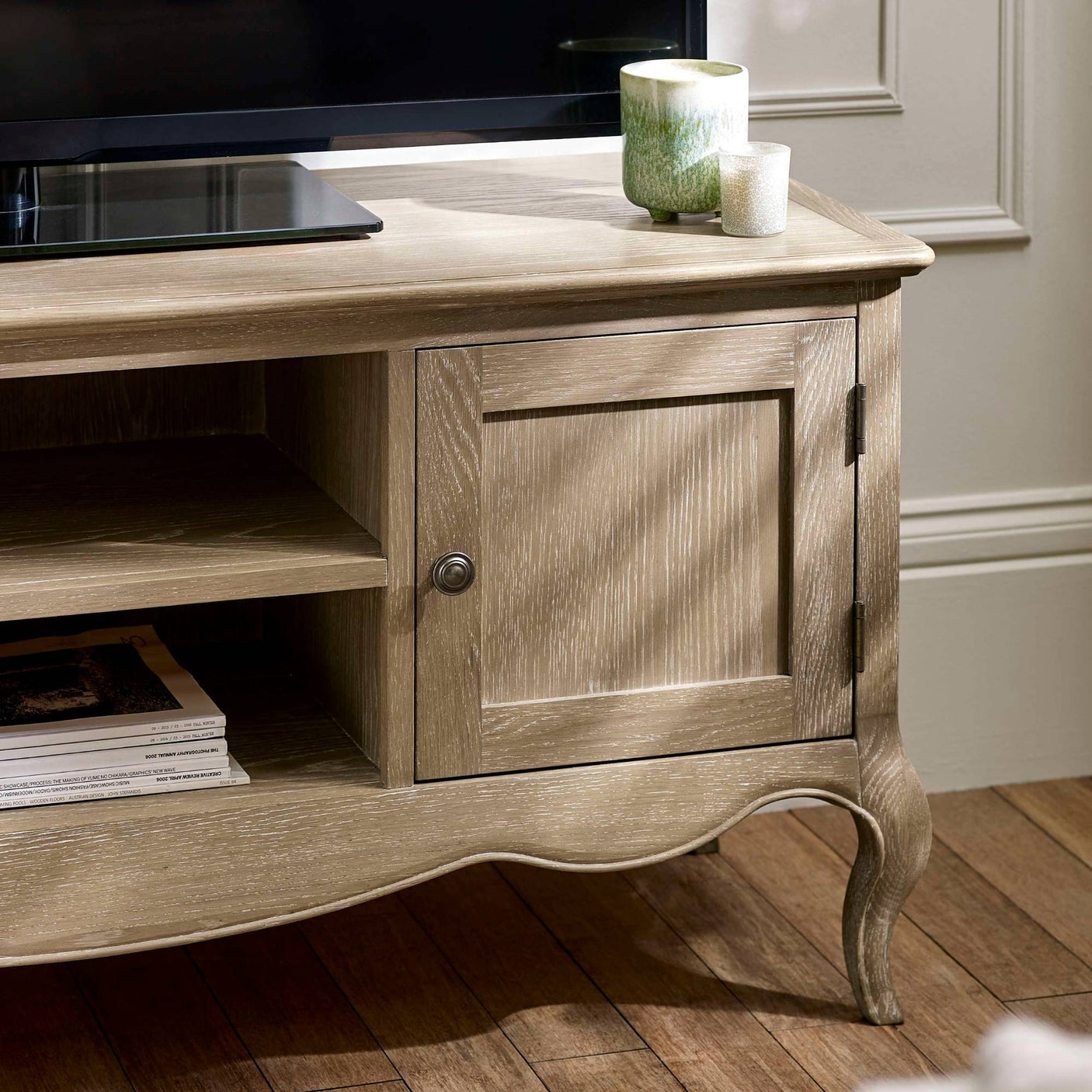 The Camille French Style TV Unit in limed oak features a textured finish with elegant curved legs, a spacious top for a television, and a combination of open shelving and a cabinet for storage, set against a softly lit room with wooden flooring