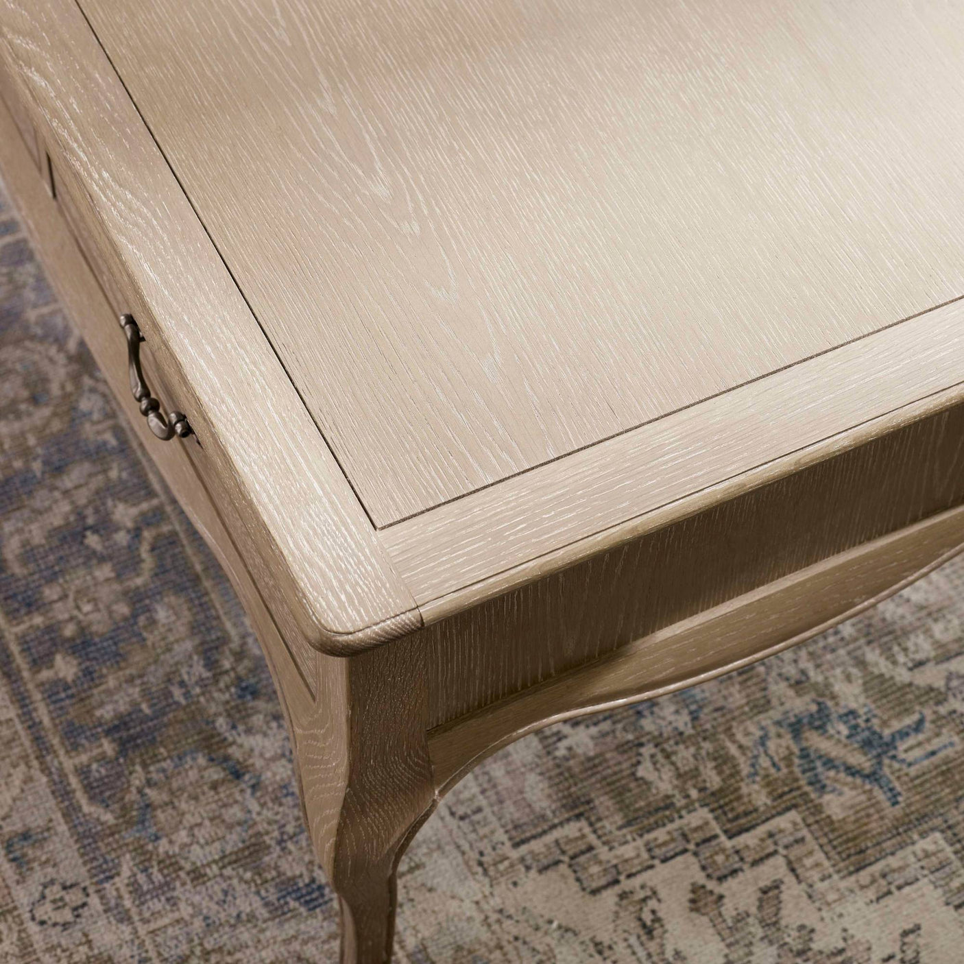The Camille French Style Rectangular Coffee Table features a limed oak finish with elegant detailing, showcasing a smooth surface and curved legs, set against a textured area rug