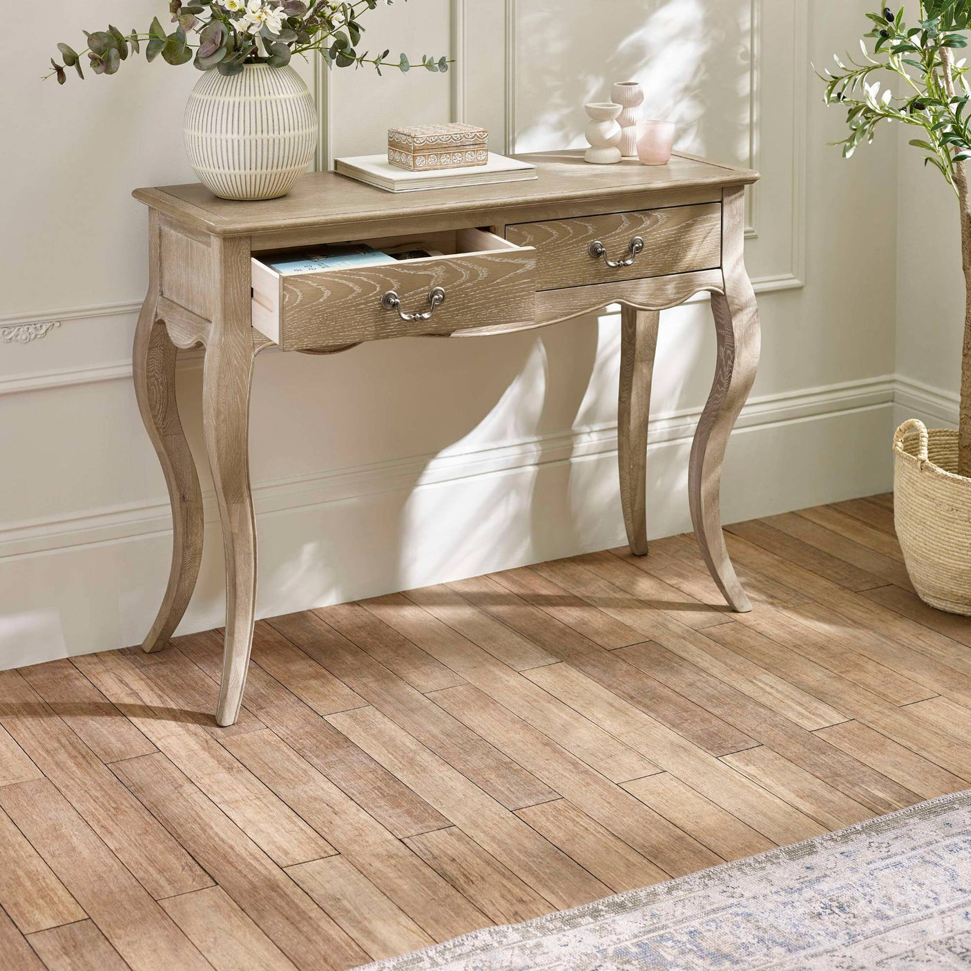 The Camille Console Table in limed oak features elegant curved legs and two drawers, set against a softly lit room with a neutral color palette and decorative accents