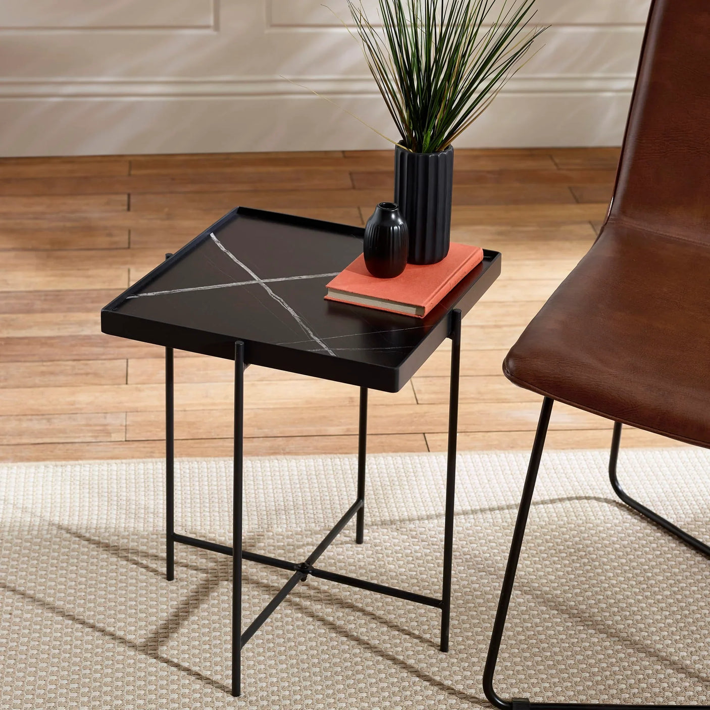 Black square side table with crossed metal legs beside brown chair on wood floor