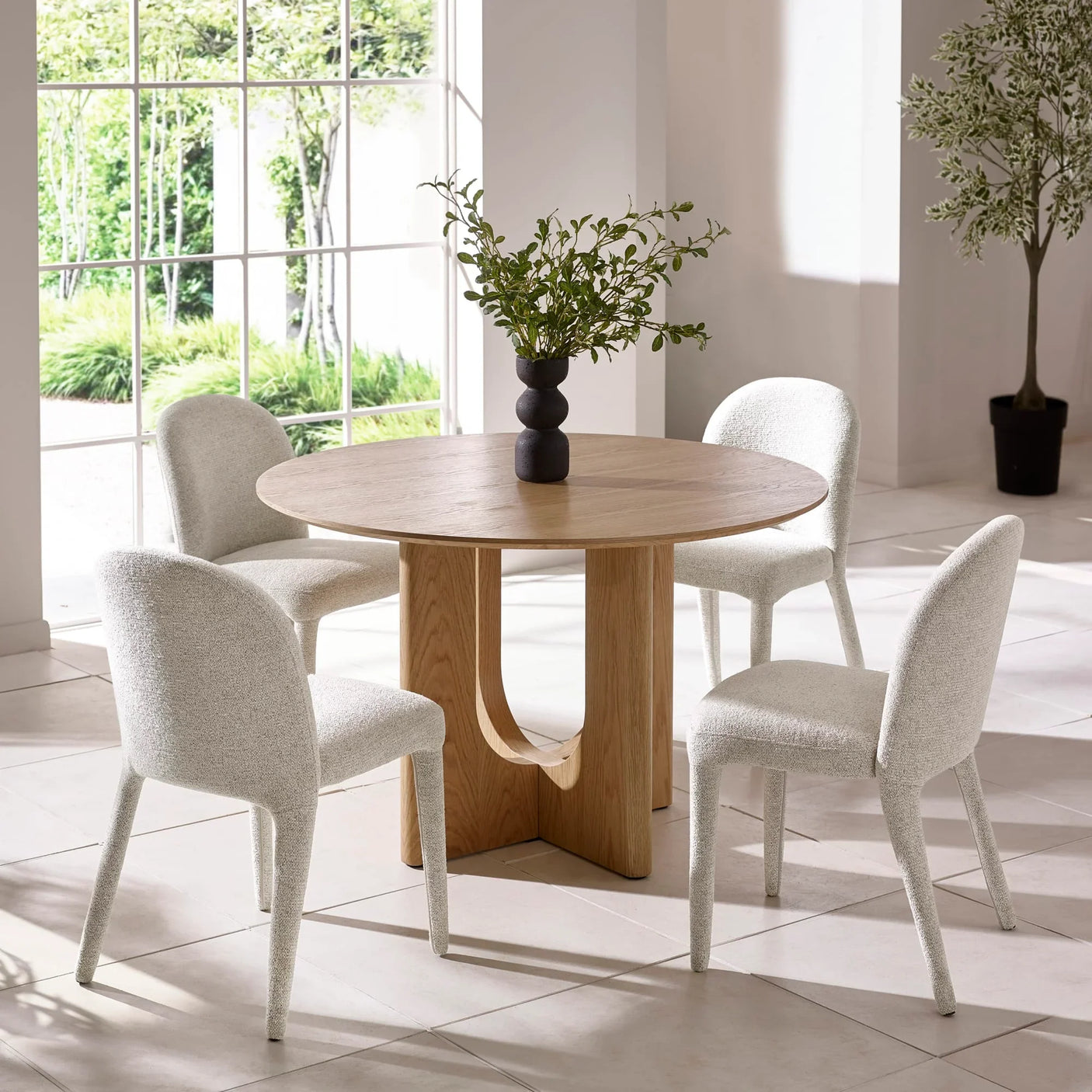 Round oak dining table with four white fabric chairs and black vase with greenery