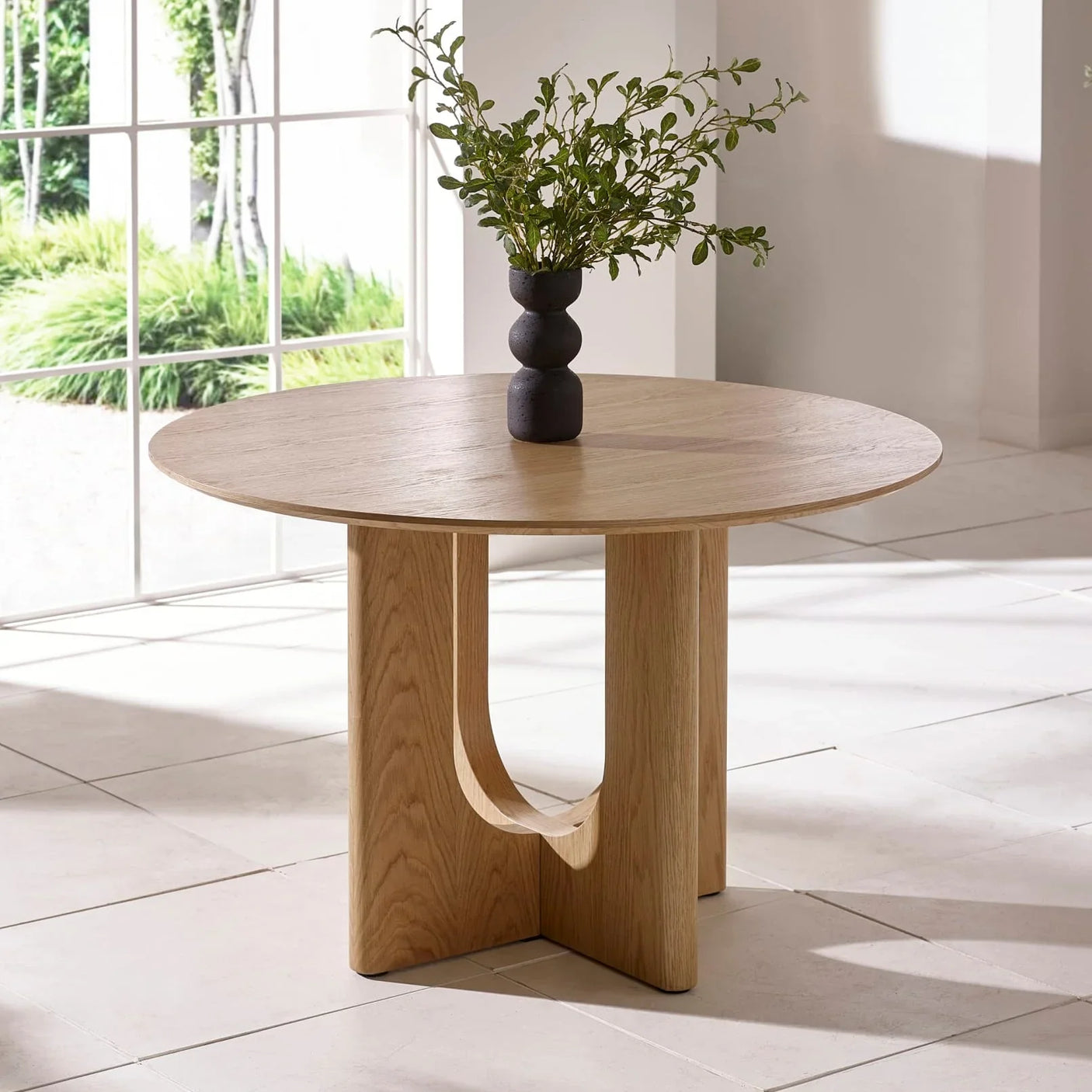 Round oak dining table with sculpted base in bright room, black vase with greenery on top