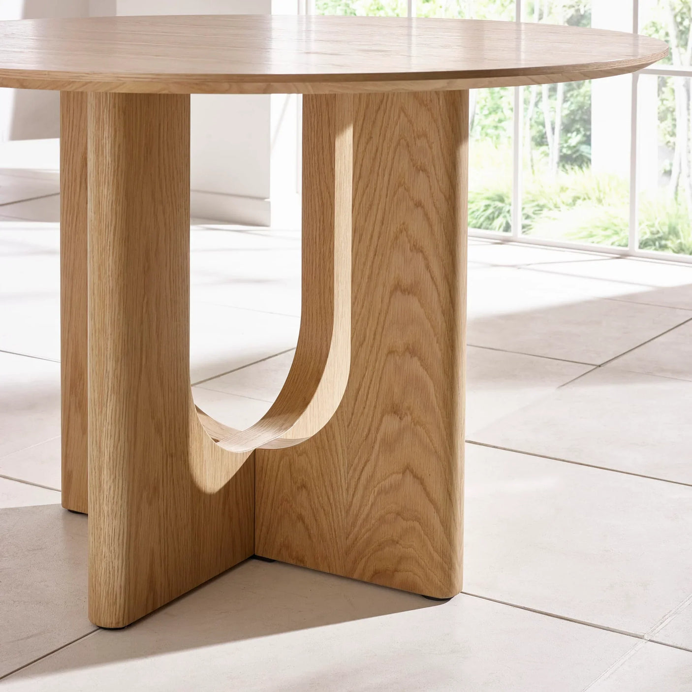 Round oak dining table with sculpted wooden base on tiled floor by window