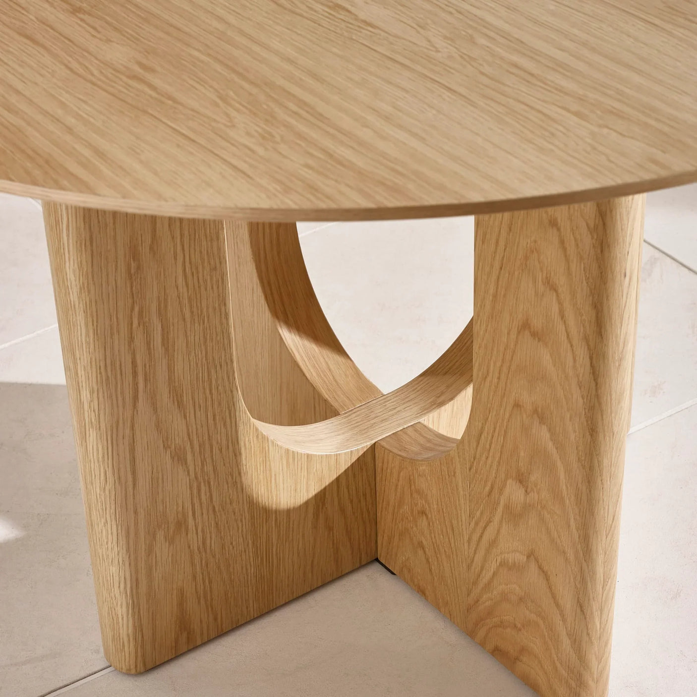 Light oak round dining table with sculpted wooden base on tiled floor