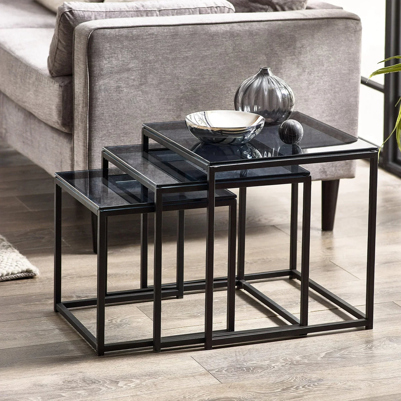 Black glass nest of tables with metal frames beside grey sofa on wooden floor
