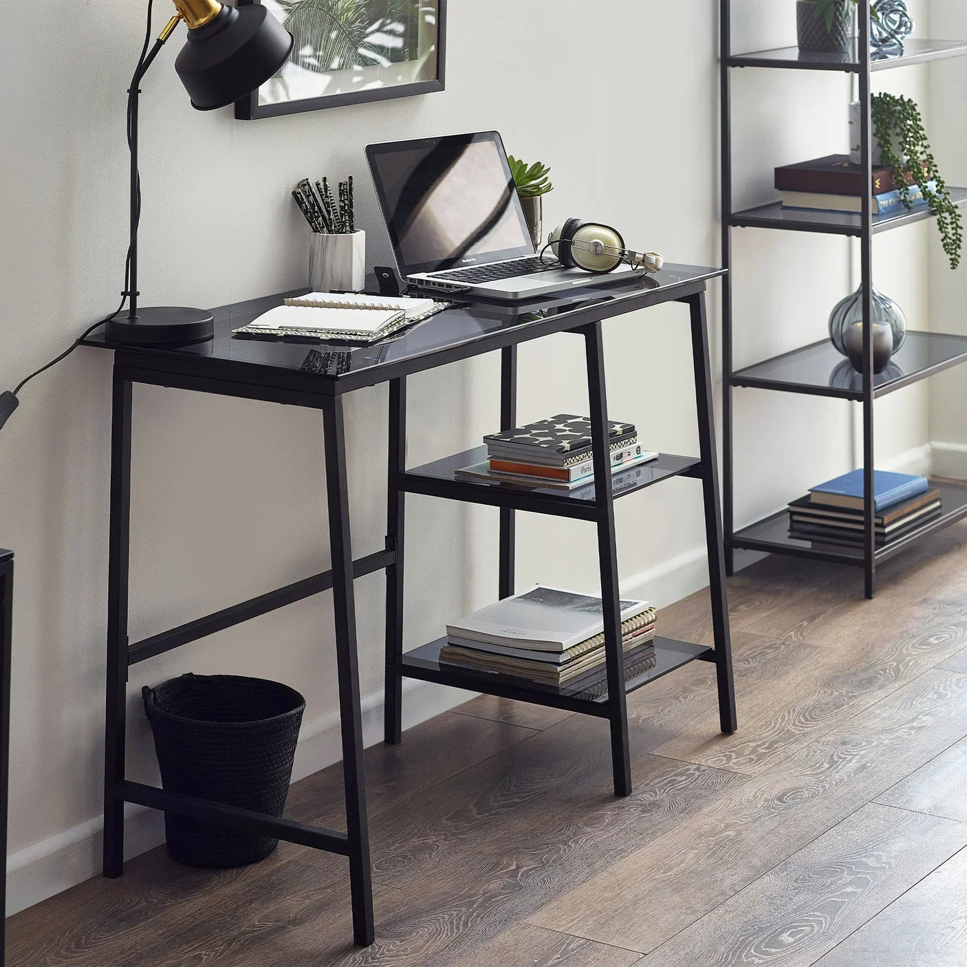Black metal desk with glass top and shelves, styled in modern home office setting