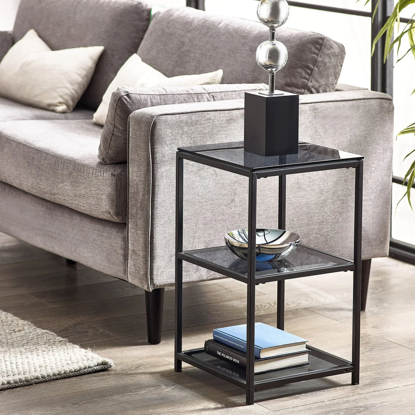 Black metal and glass tall narrow side table beside grey sofa with books and decor items