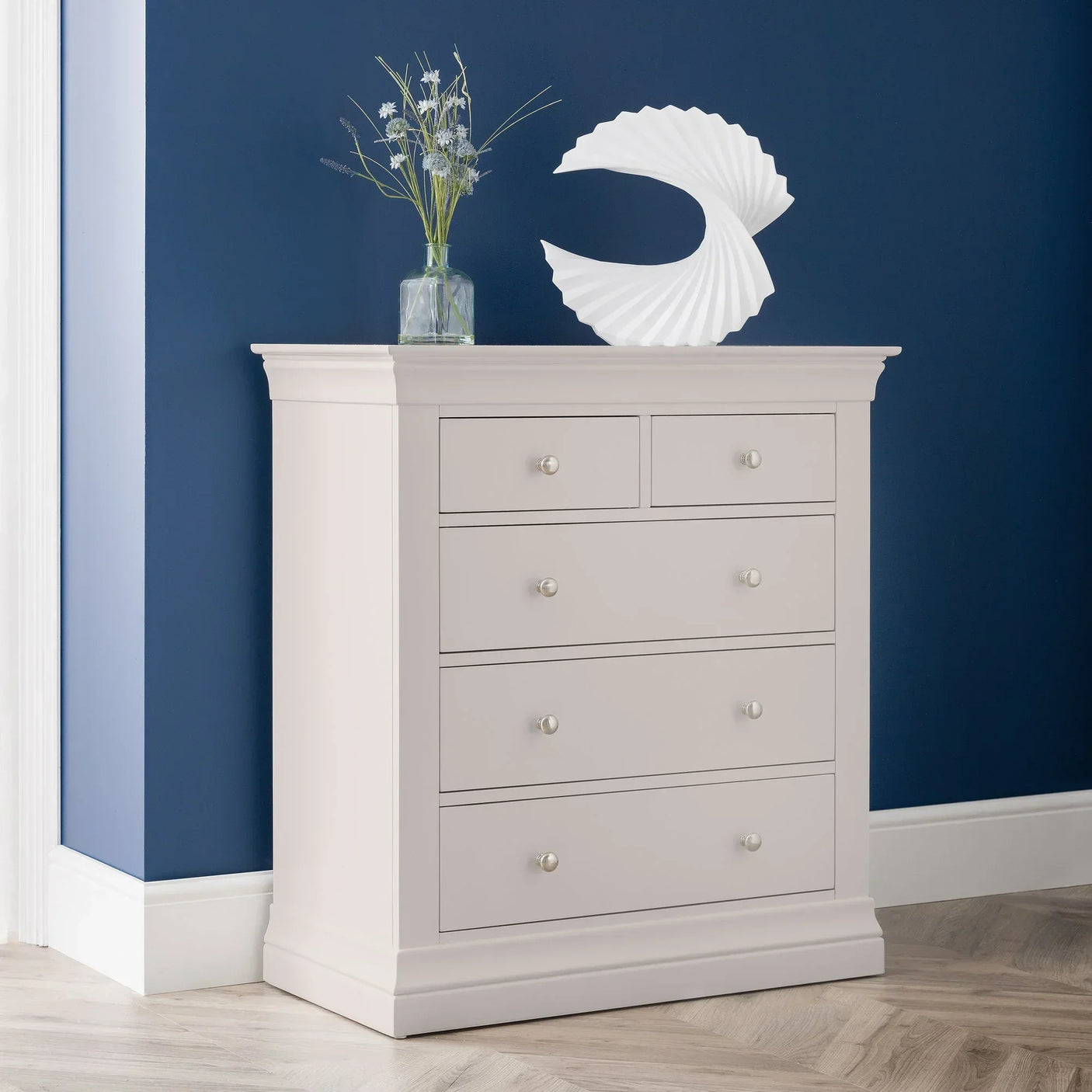 Light grey chest of drawers with round knobs, styled against blue wall with vase and sculpture