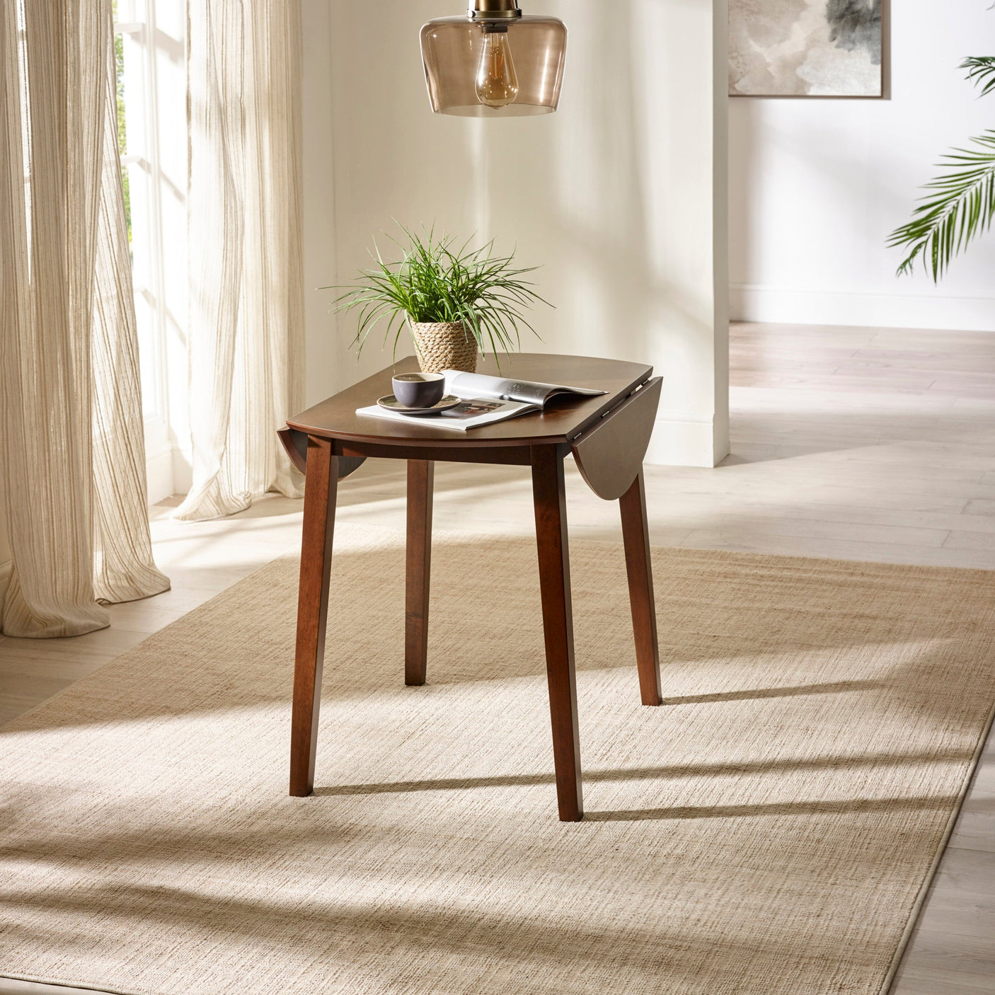 Brown wooden round table with one drop leaf open, angled legs, beige rug and potted plant on top