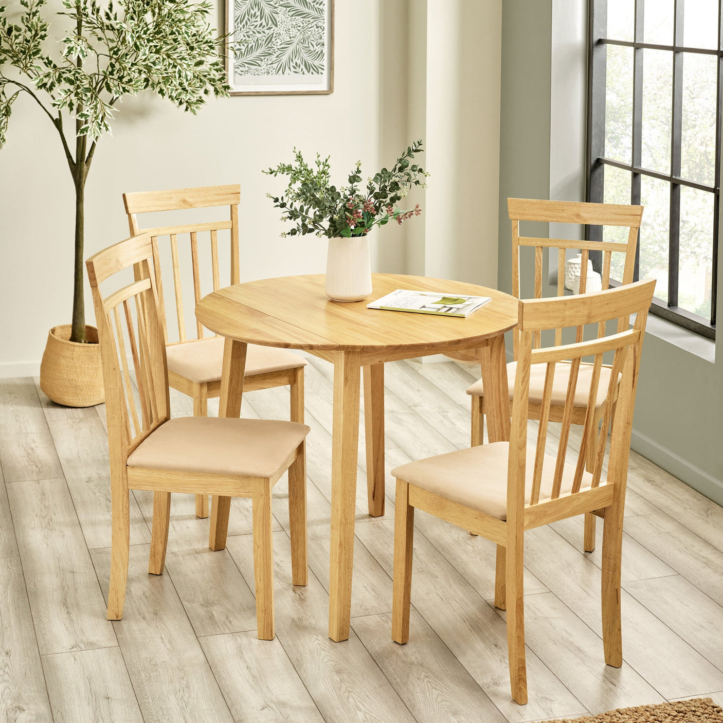 Four light wood dining chairs with beige padded seats around round table in a bright room with window and plant