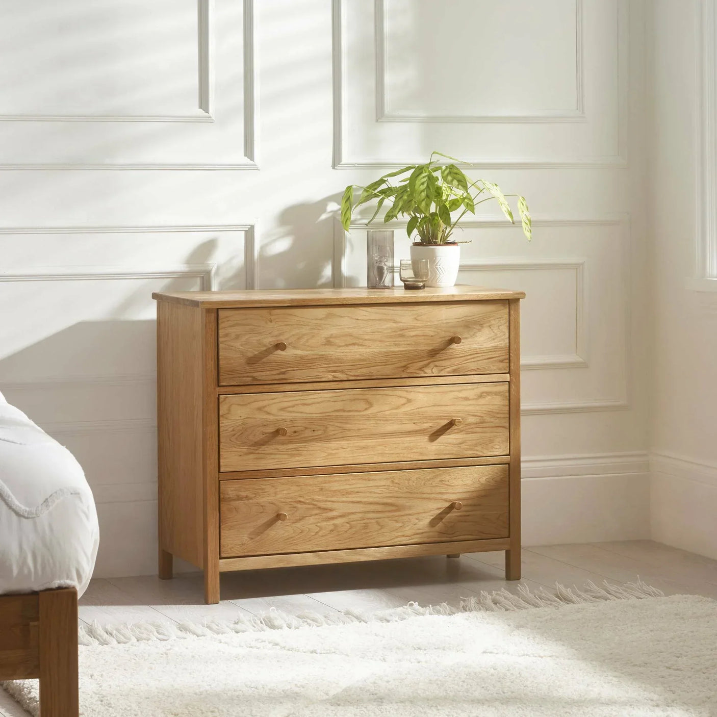 Oak 3 drawer chest with wooden handles in bright bedroom, plant and decor on top