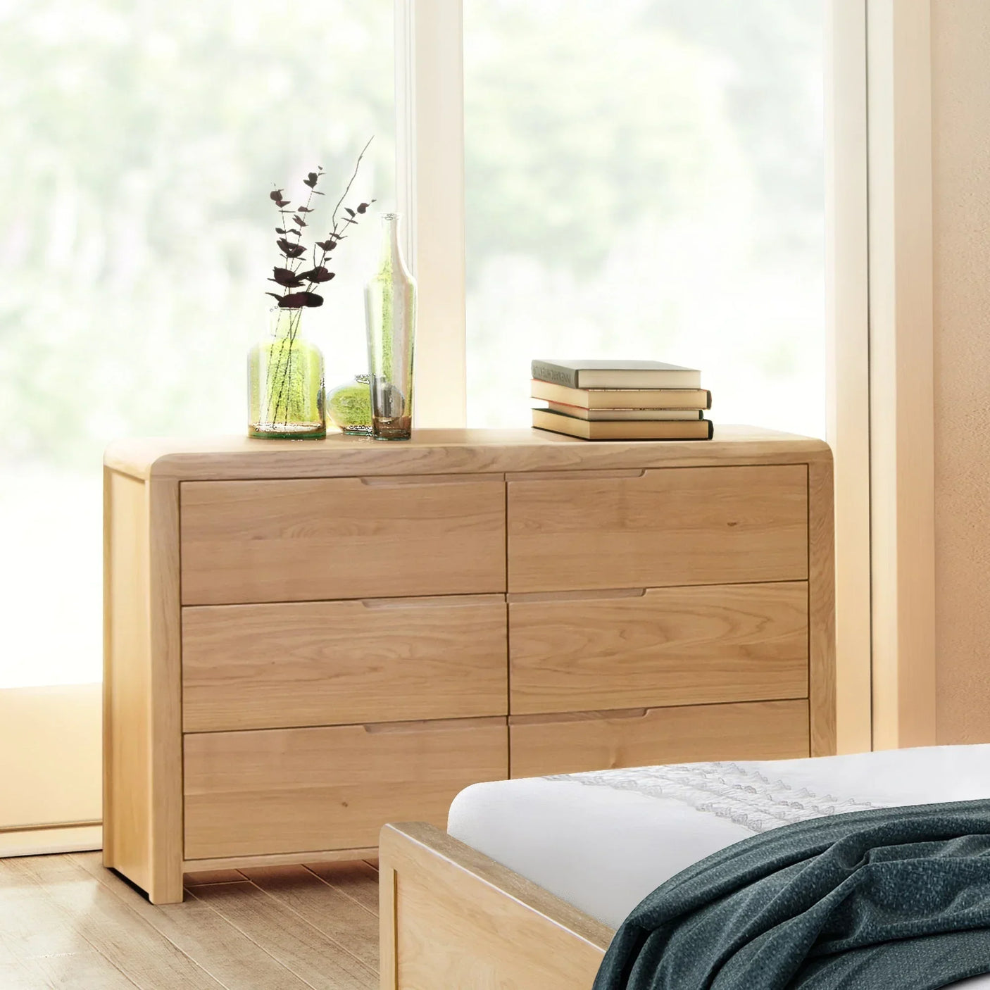 Natural wood 6 drawer wide chest in bedroom with glass vases and books on top