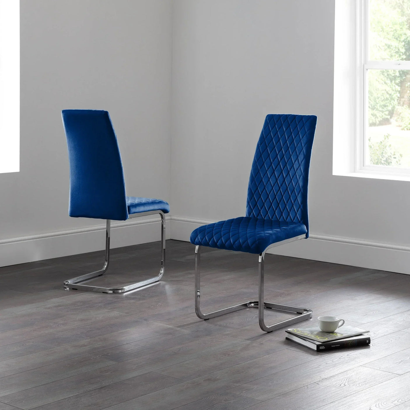 Blue velvet cantilever dining chairs with chrome legs on dark wood floor near window