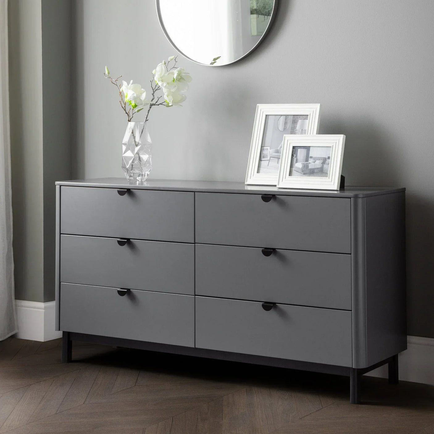Grey 6 drawer chest with black handles, vase, and photo frames in modern bedroom setting