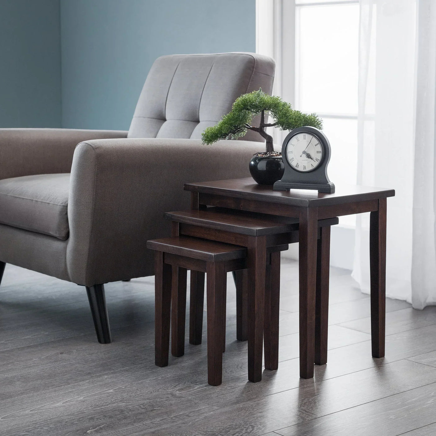 Mahogany nest of tables set beside grey armchair with clock and bonsai plant on tabletop