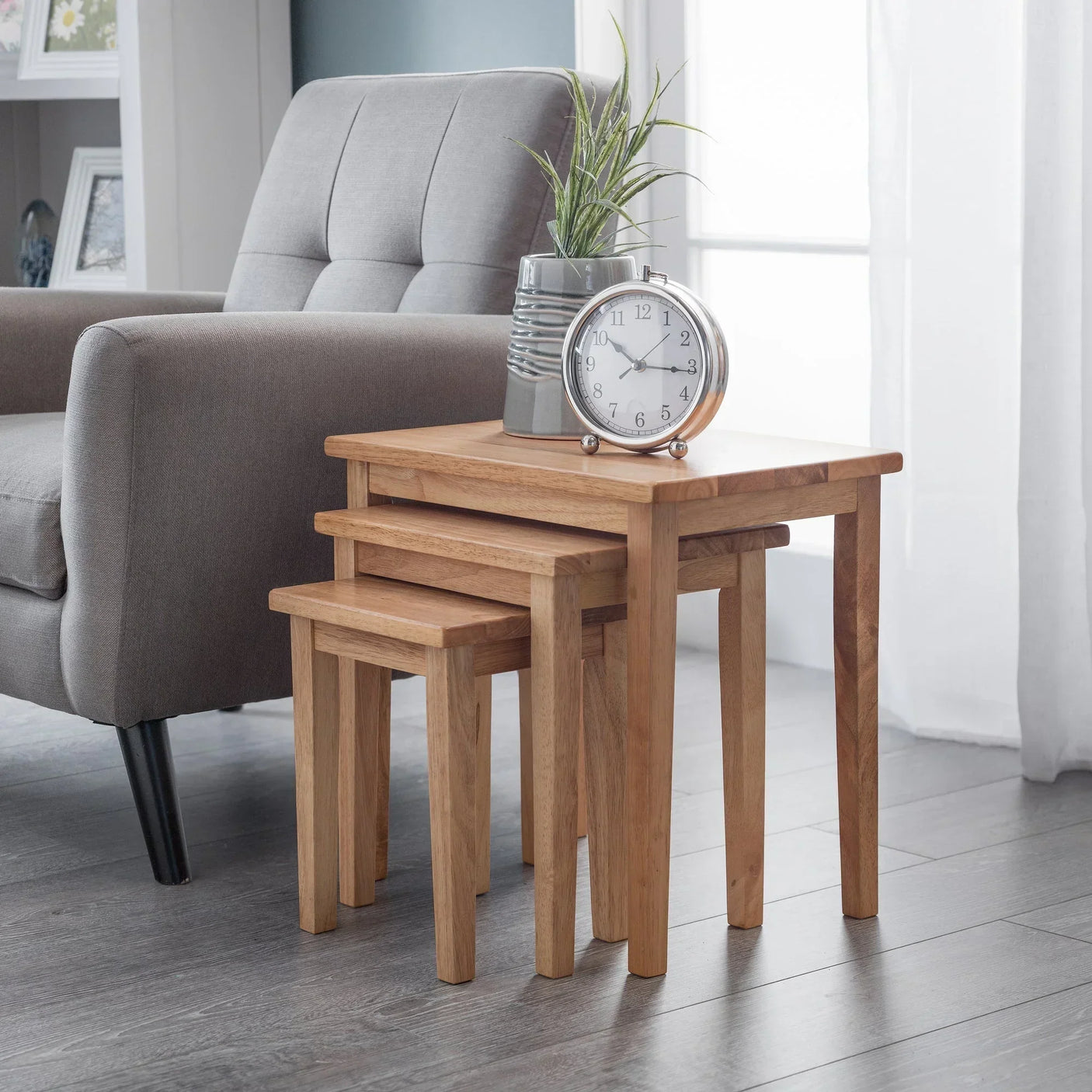 Oak nest of tables set beside grey armchair with clock and plant decor