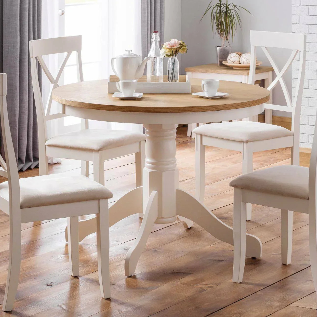 Round pedestal dining table with light wood top and four white chairs in a bright room