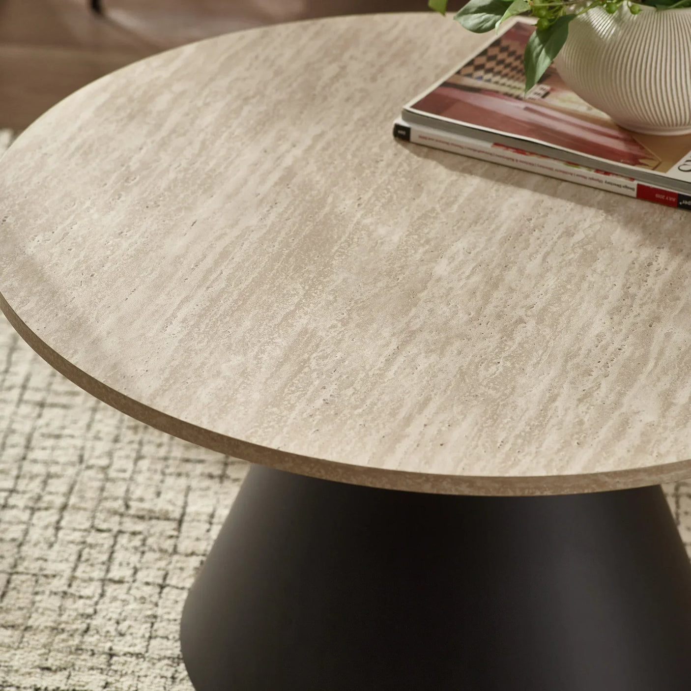 Round coffee table with light stone top, black base, books and plant on textured rug