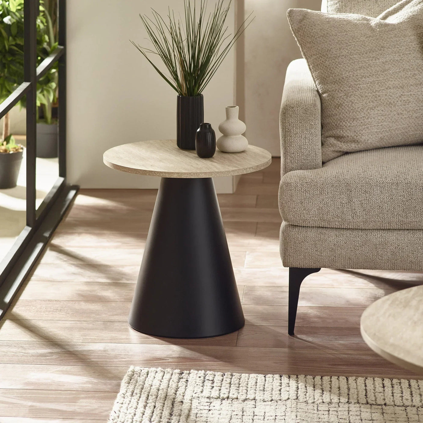 Round side table with black cone base and light wood top beside beige sofa in living room