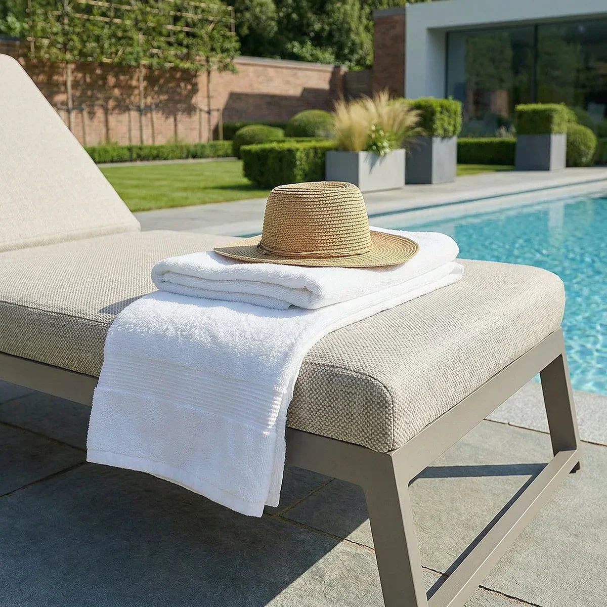 Aluminium sun lounger with beige cushion by pool, topped with white towels and straw hat