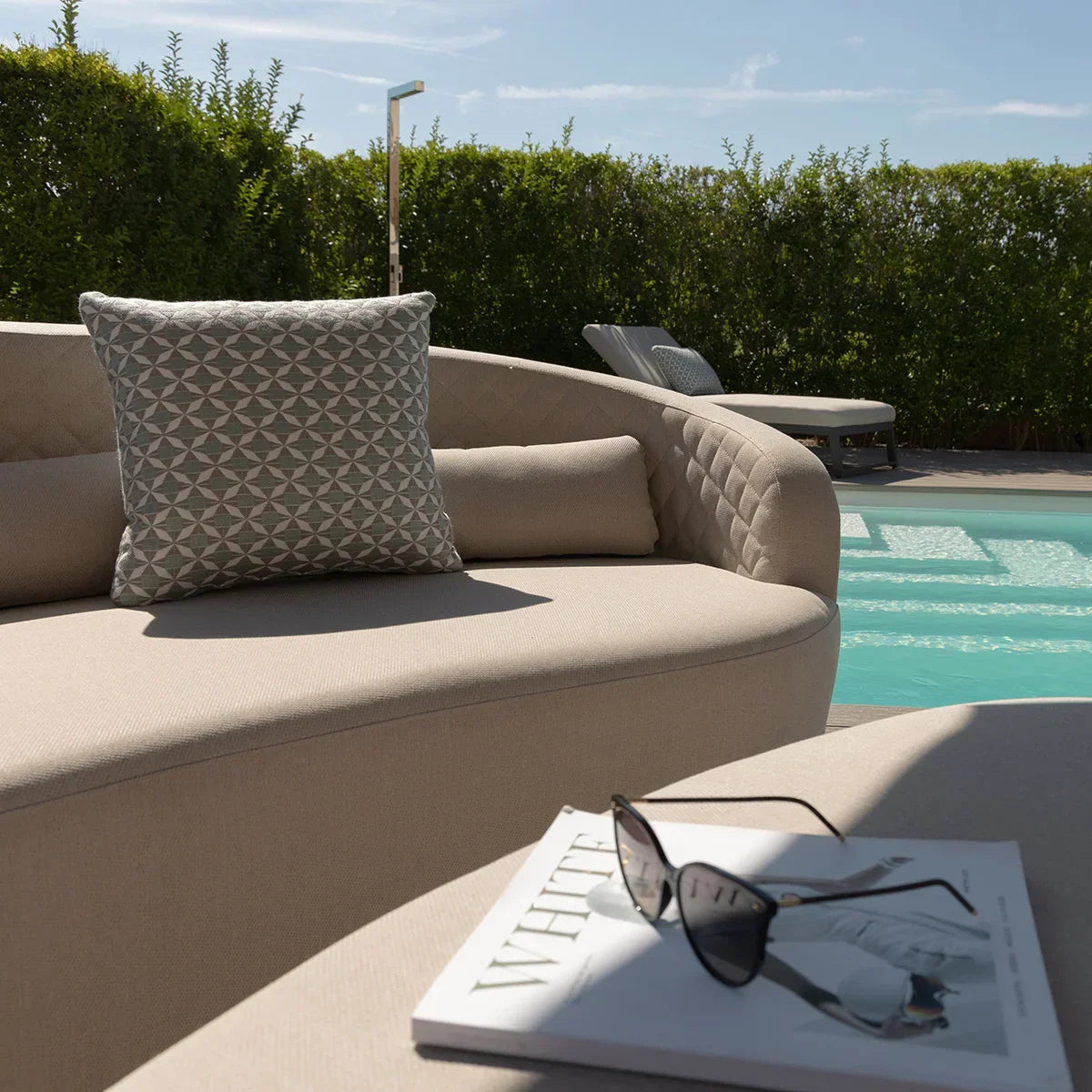 Outdoor beige fabric sofa with patterned cushion by pool, sunglasses and magazine on ottoman