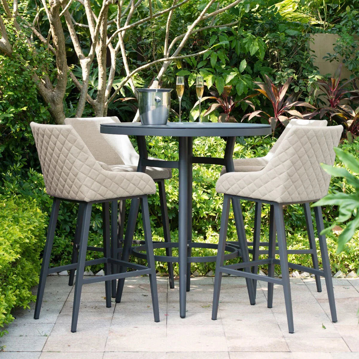 Outdoor bar table and four beige quilted bar stools on patio surrounded by greenery