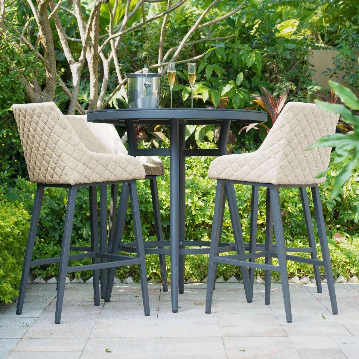 Outdoor bar table set with two quilted beige bar stools on a stone patio in garden setting
