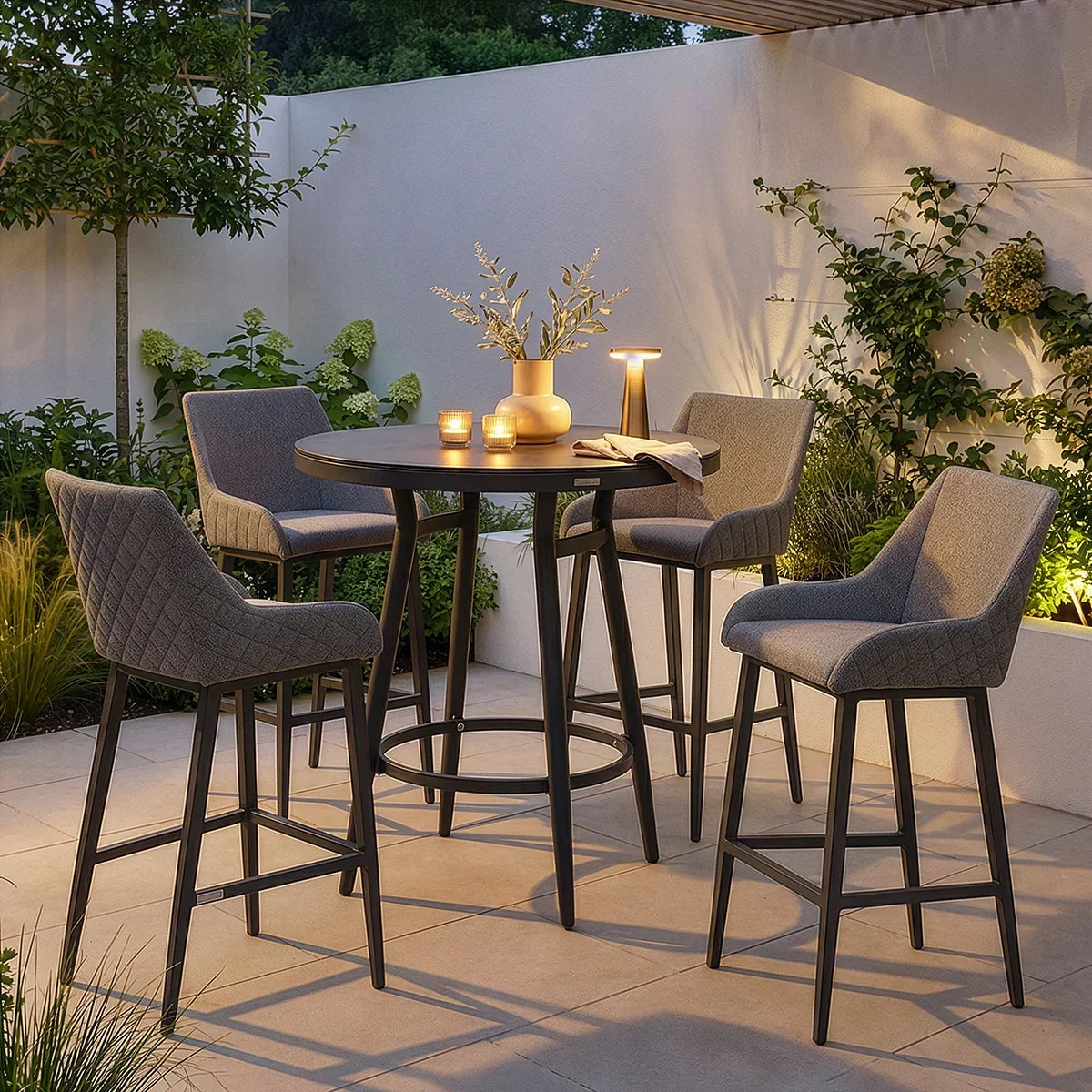 Grey outdoor bar table set with four cushioned bar stools on a tiled patio at dusk