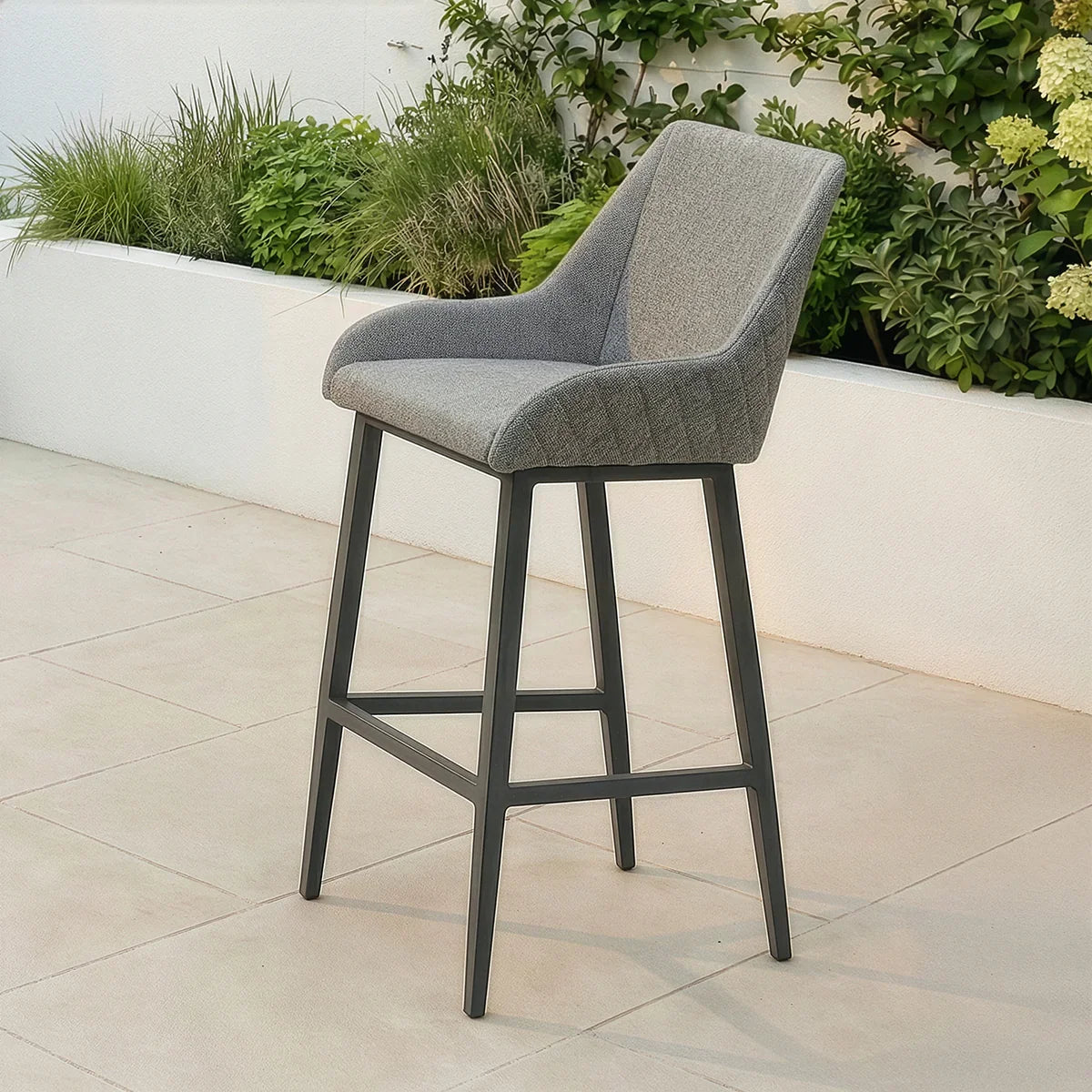 Grey bar stool with upholstered seat and black metal legs on tiled patio near greenery
