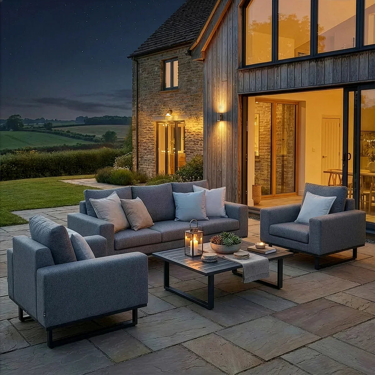 Grey outdoor sofa set with armchairs and cushions on stone patio, modern house in background