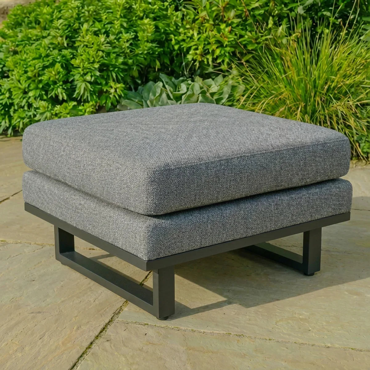 Grey outdoor footstool with cushion and metal frame on patio with plants in background