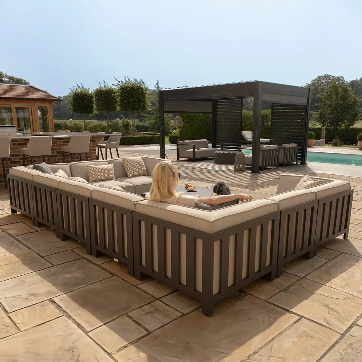 Large outdoor corner sofa set with beige cushions on patio beside garden pergola and pool