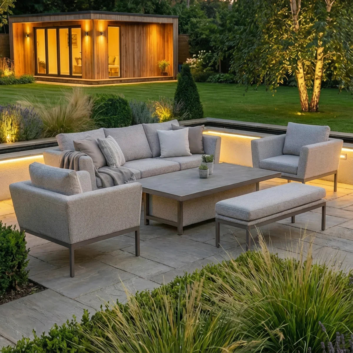 Grey outdoor sofa set with armchairs, bench and table on a stone patio in landscaped garden