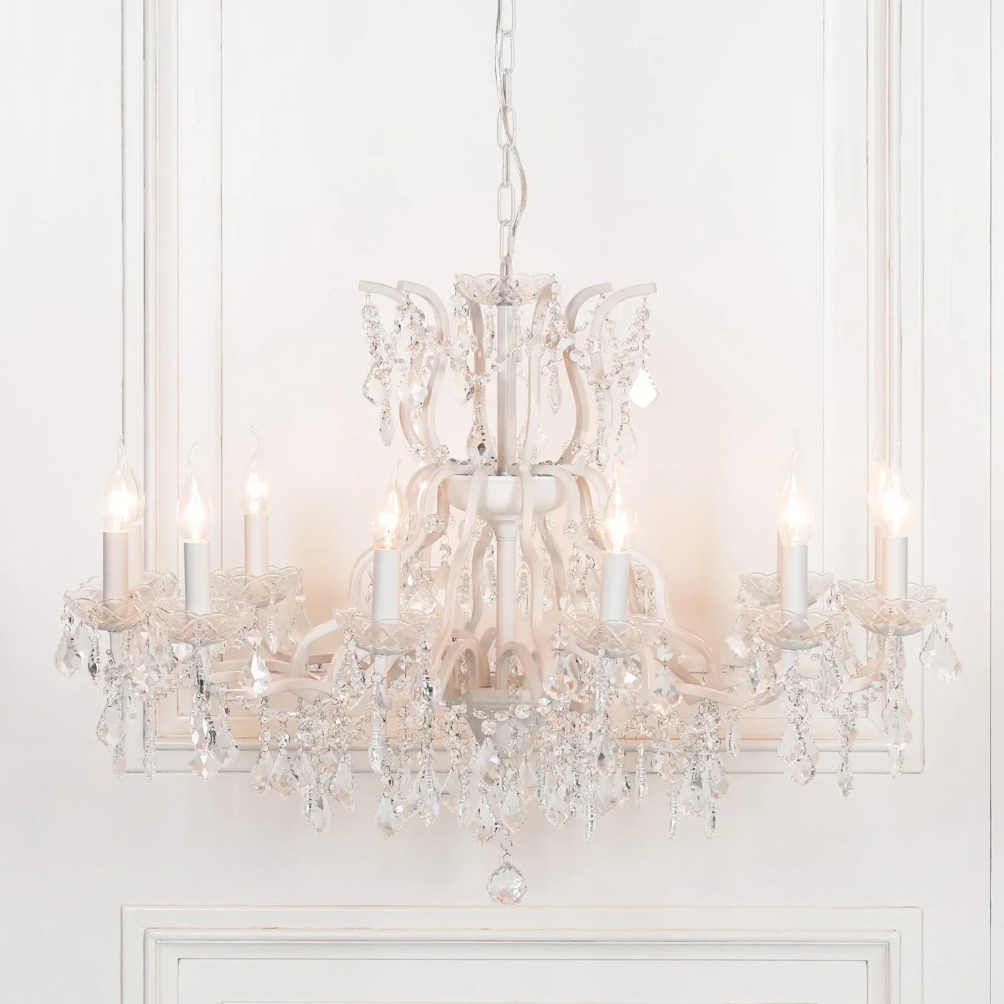 White crystal chandelier with candle-style lights and hanging glass droplets against white wall
