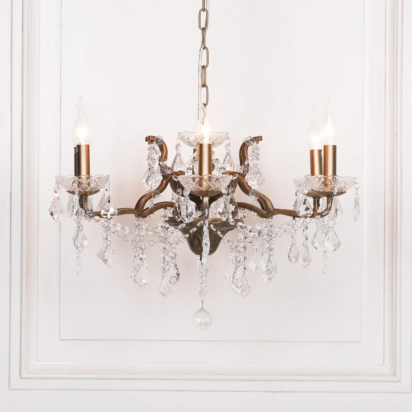 Crystal chandelier with gold finish and candle-style bulbs hanging against white panelled wall