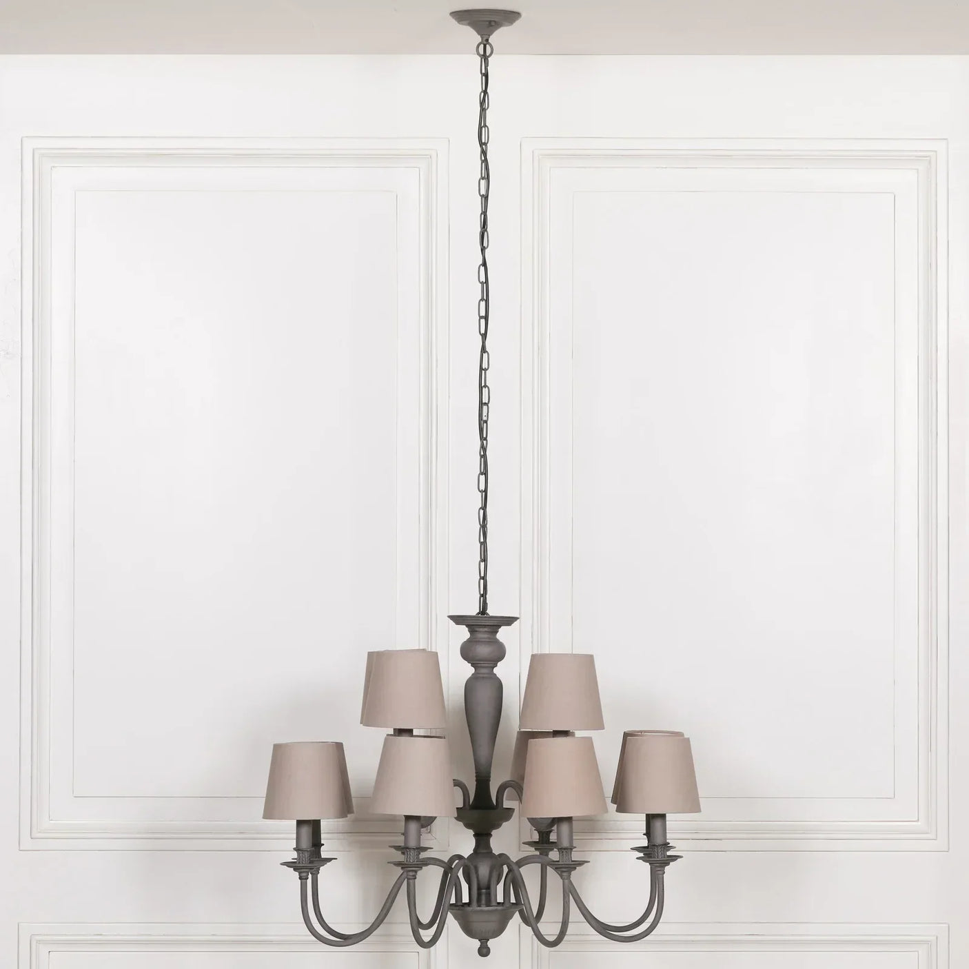 Grey six arm chandelier with taupe fabric shades hanging in front of white paneled wall
