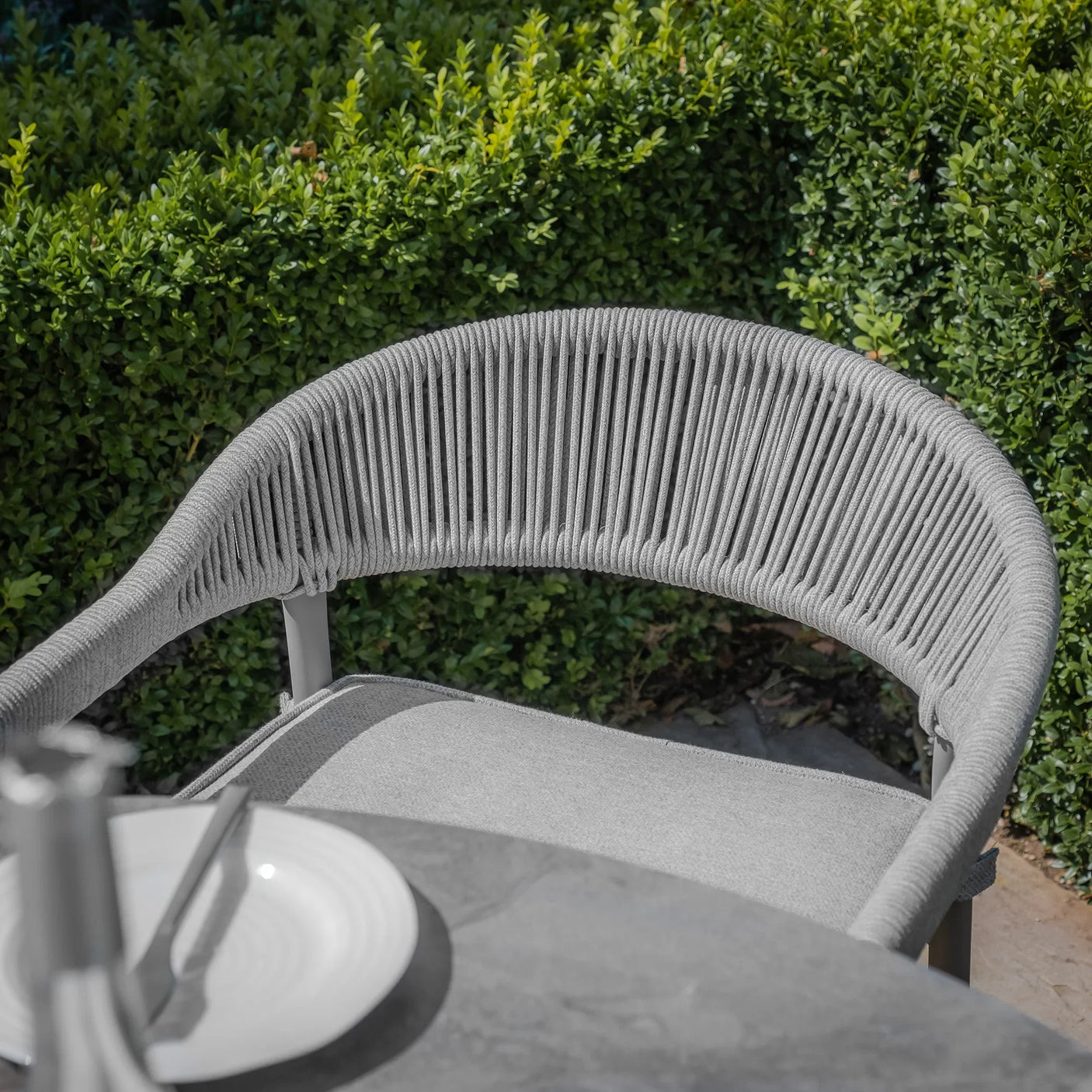 Grey outdoor rope dining chair with curved backrest beside patio table and greenery