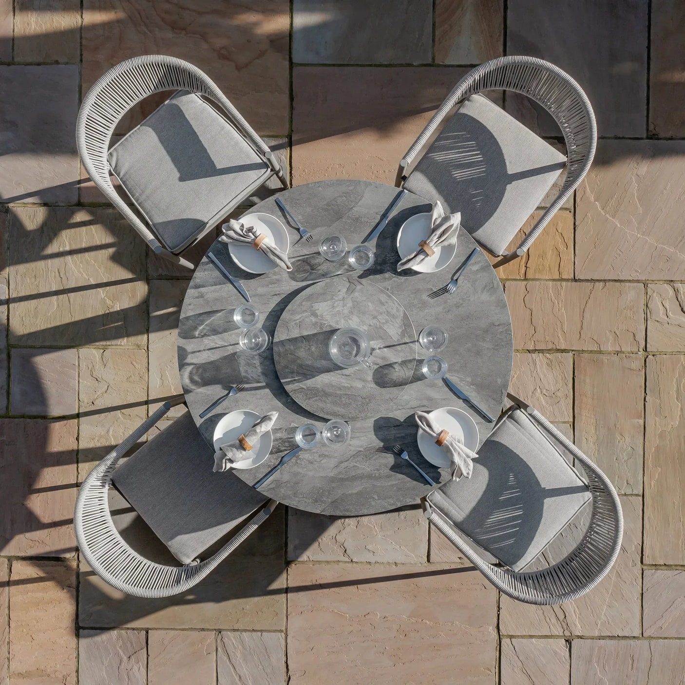 Round grey stone dining table set with four cushioned chairs and tableware on patio