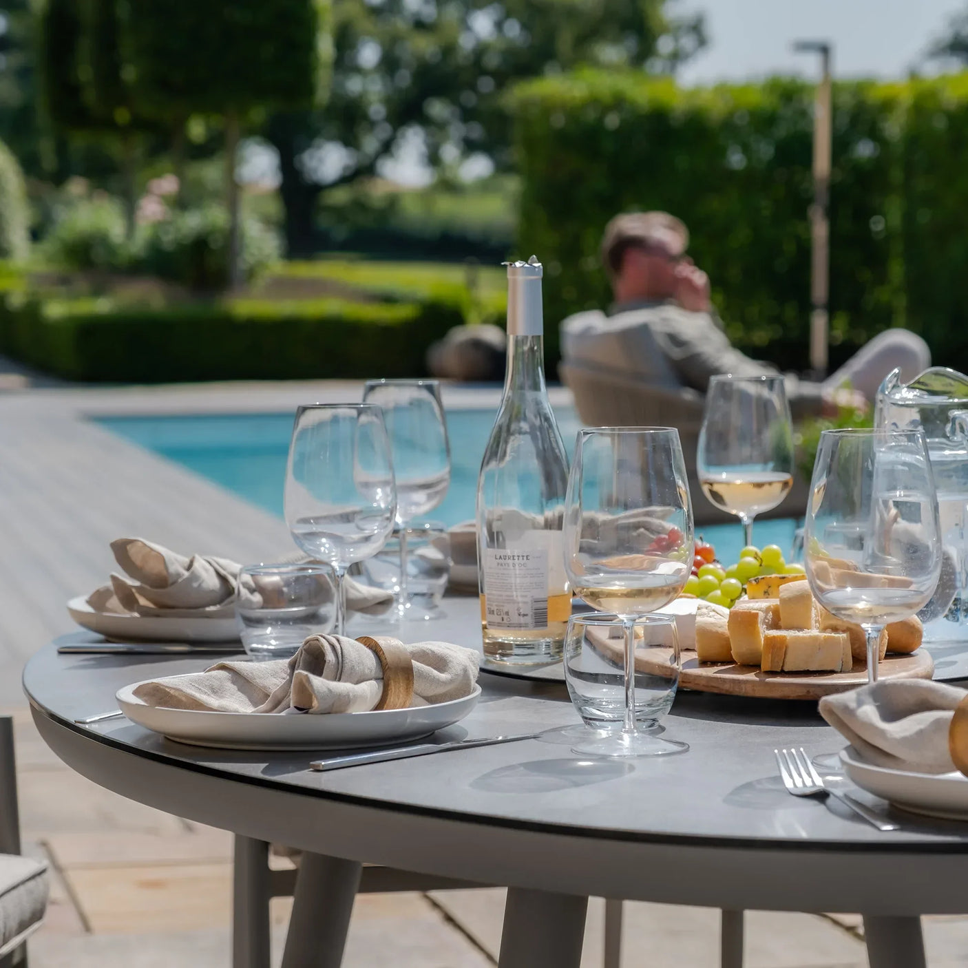 outdoor dining table set with plates, glassware, bread and fruit beside a swimming pool