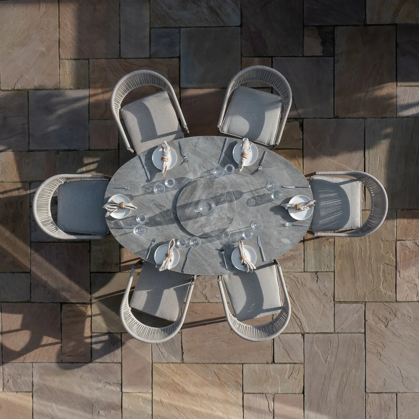 Grey outdoor dining set with six cushioned chairs and oval stone table on patio tiles