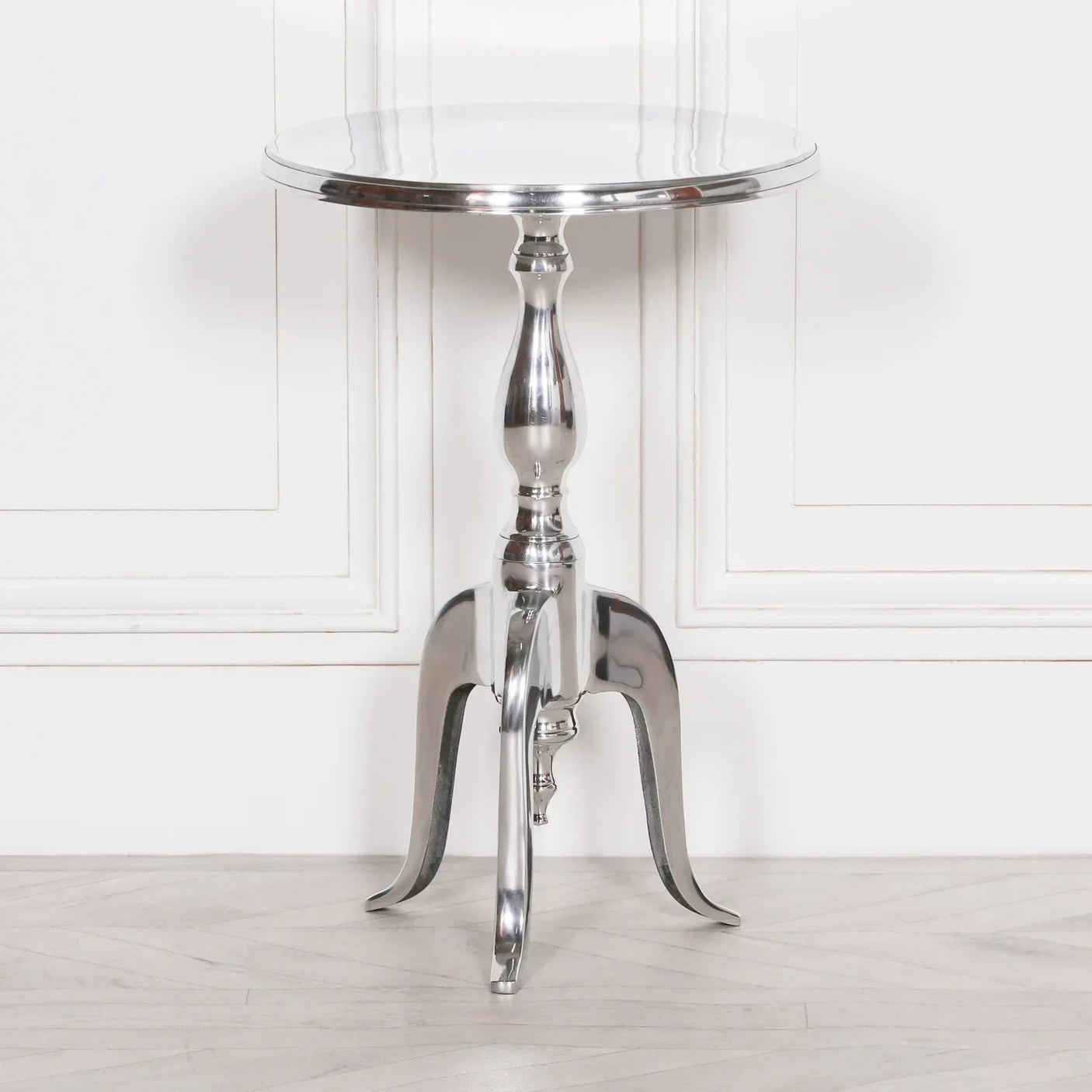 Round silver metal side table with tripod legs and polished finish against white panel wall