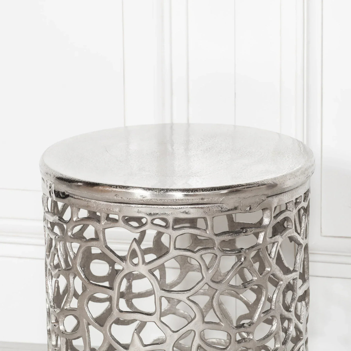 Silver metal side table with openwork design and round top against white panel wall