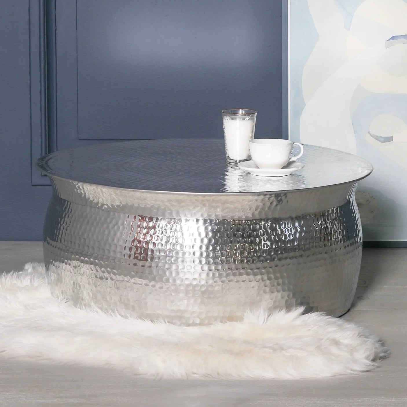 Round hammered silver coffee table with candle and cup on top, set on white faux fur rug