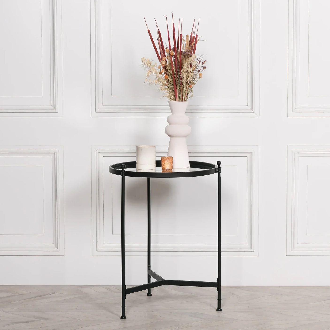 Black metal side table with round top, vase of dried flowers and candles against white wall