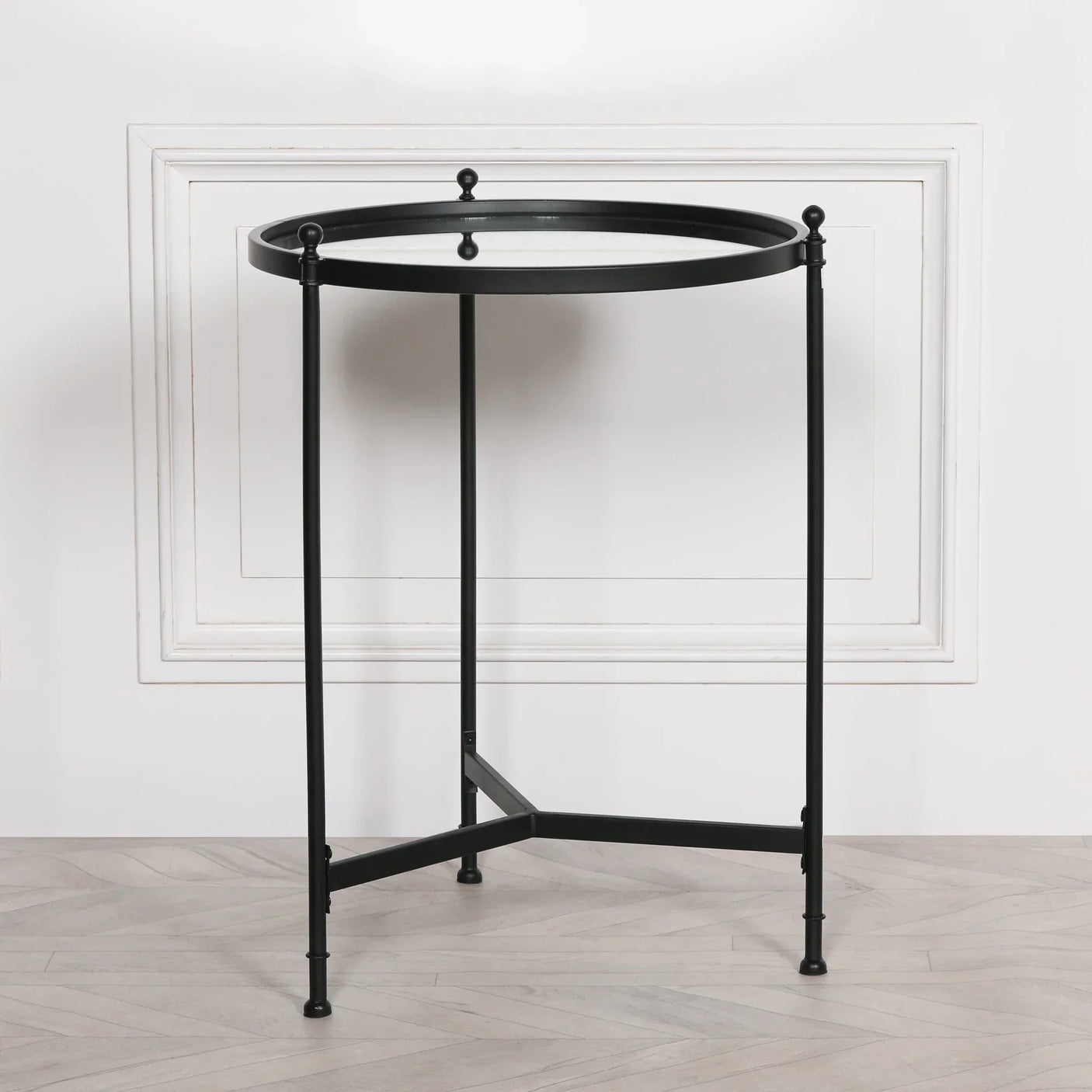 Black metal side table with round glass top and slim legs on light wood floor