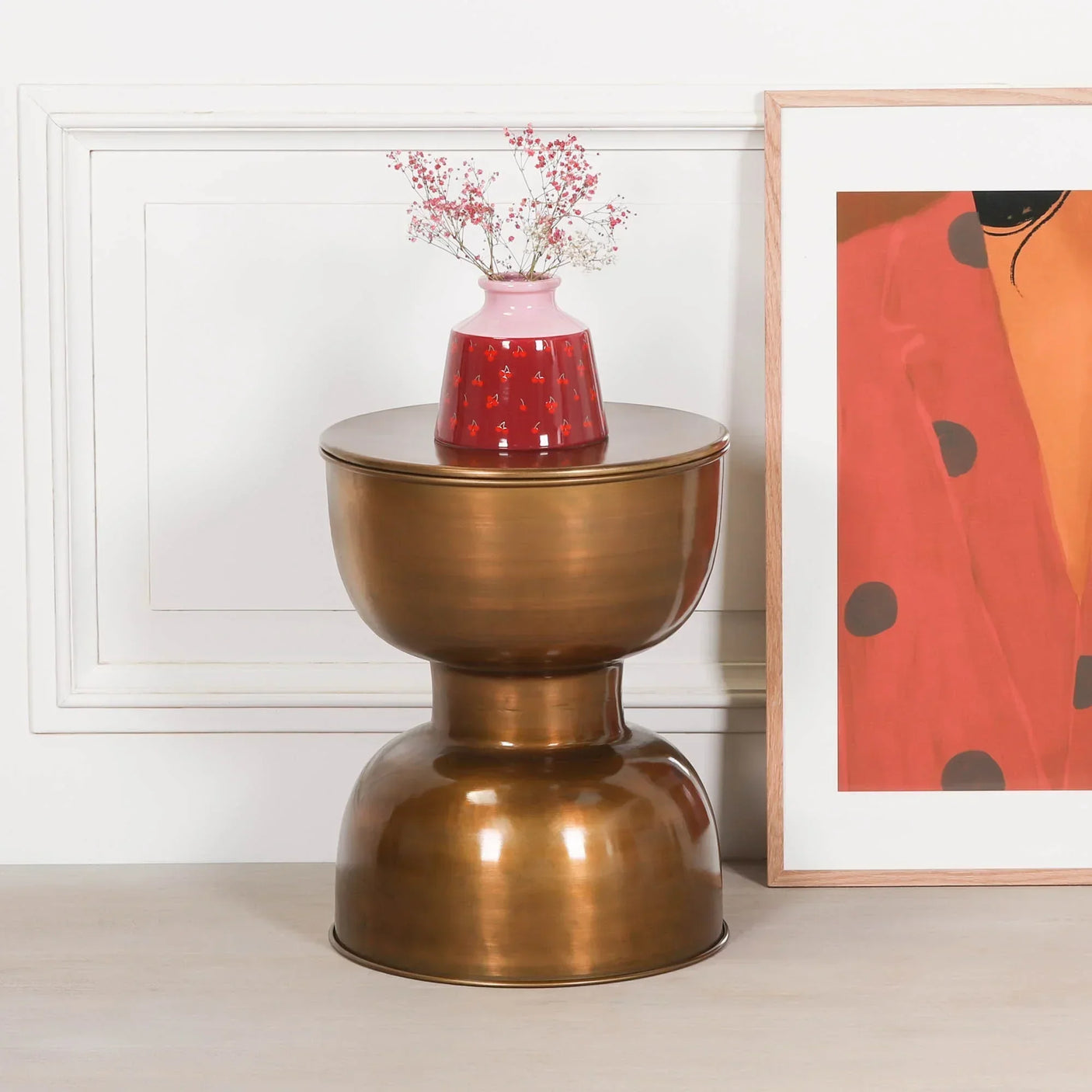 Brushed brass side table with rounded base, pink and red vase on top, framed art beside