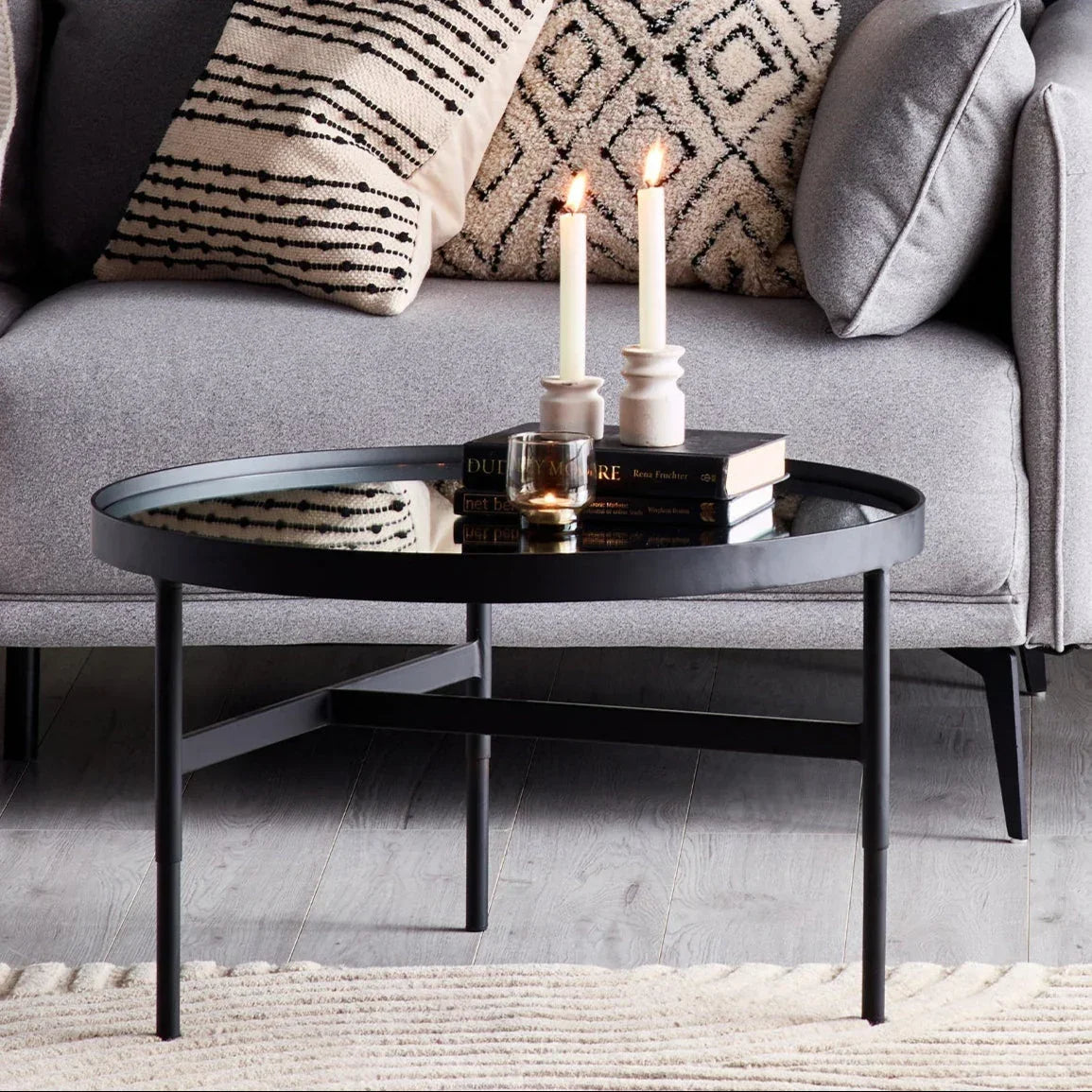 Black metal coffee table with glass top, two candles, books, grey sofa and patterned cushions